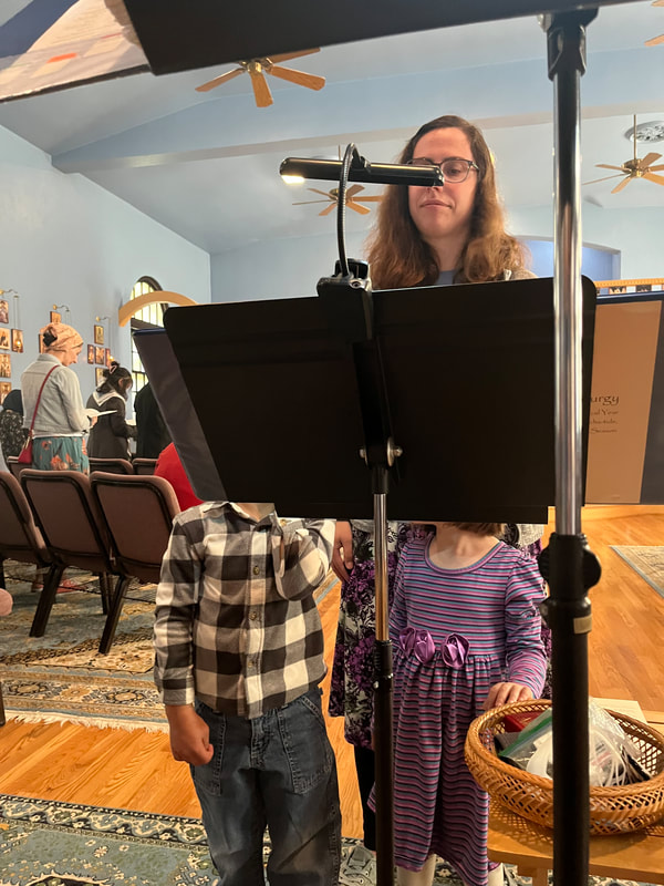A woman stands behind a choir stand. Two small children stand in front of her. Their faces are blocked by the music stand.