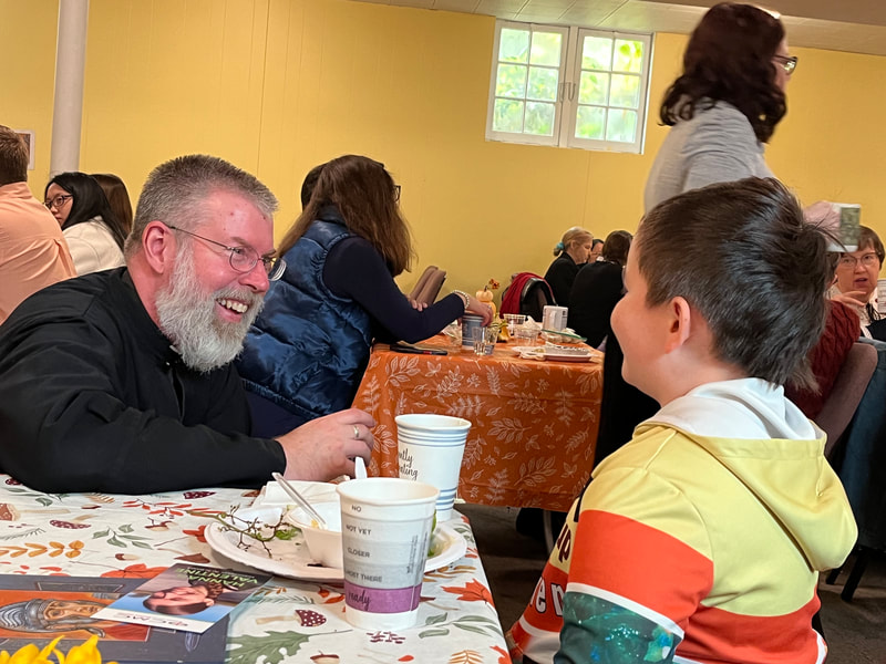 A priest with a beard and a young boy sit together at a table. They are smiling and conversing. The tables around them are full of people.