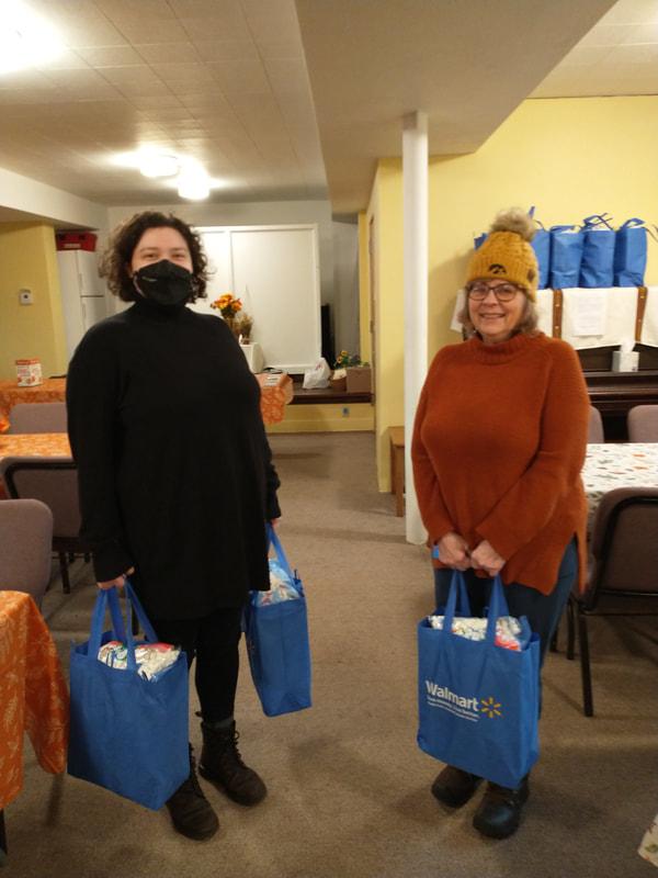 Two women holding blue reusable grocery bags filled with holiday food items.