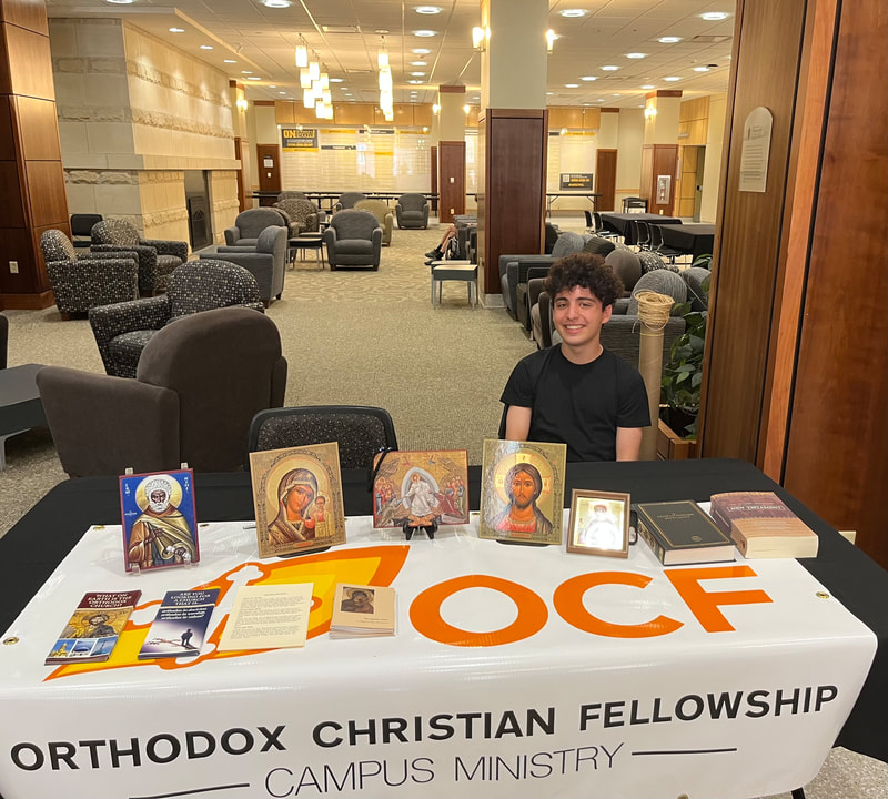 A young man smiles at the camera. He is seated behind a table covered with a banner reading "OCF, Orthodox Christian Fellowship." On top of the table are books, brochures, and icons.